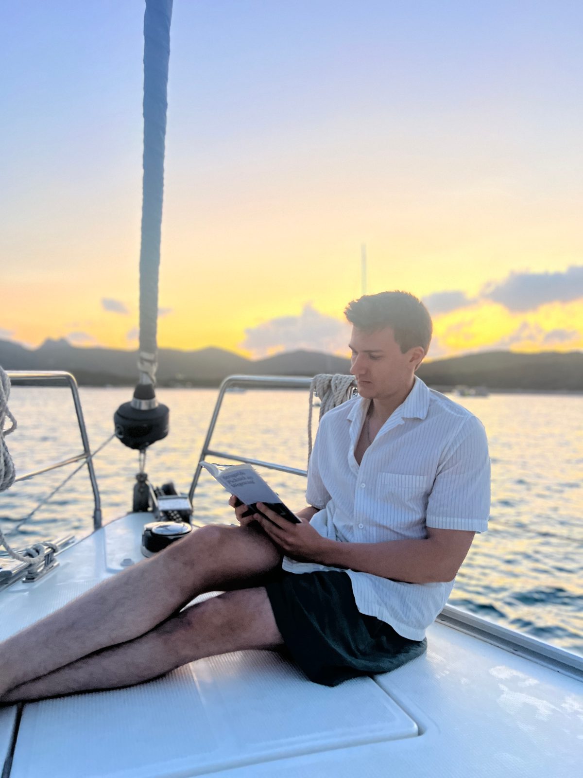 Dominik Jäkel liest an Deck eines Segelboots, im Hintergrund Berge im Abendlicht